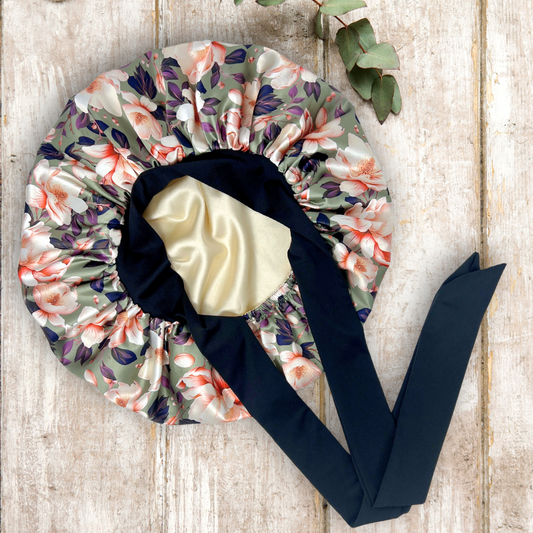 Elegant olive colored satin bonnet with a floral print, featuring a black stretch tie band for a secure and stylish fit, displayed on a wooden background.