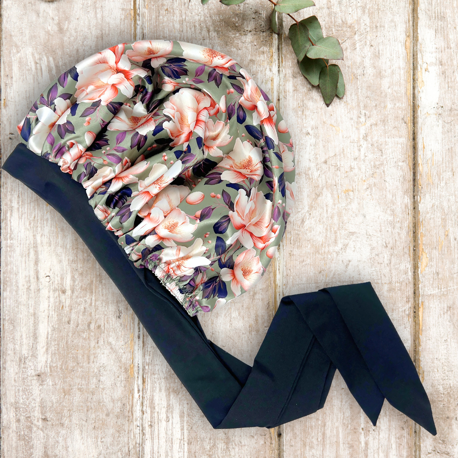 Elegant olive colored satin bonnet with a floral print, featuring a black stretch tie band for a secure and stylish fit, displayed on a wooden background.