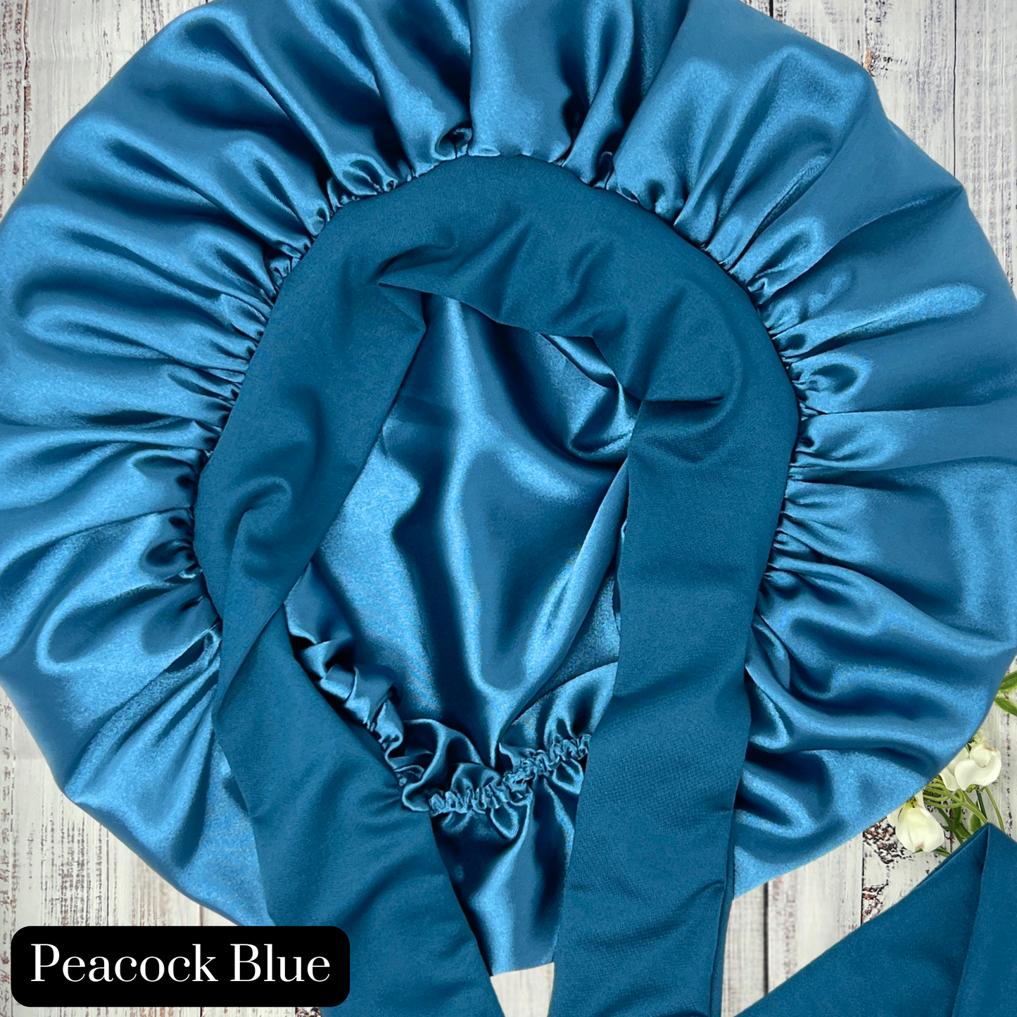 Close up view of a Peacock blue satin hair bonnet displayed on a light wood background, showcasing smooth satin texture and vibrant color. Ideal for curly, natural, and protective hairstyles.