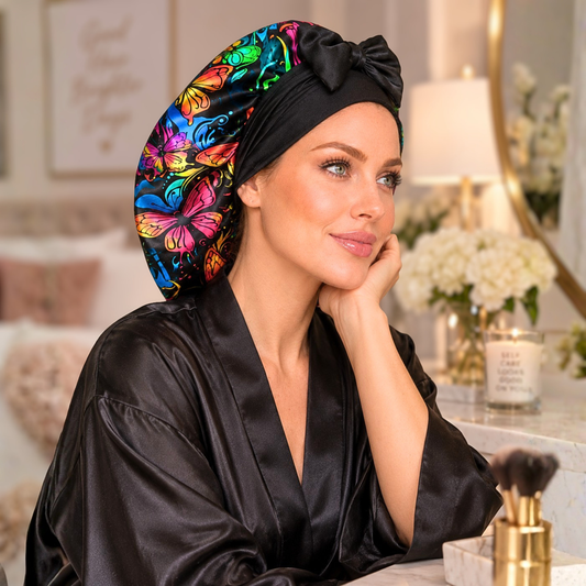 A woman wearing a black satin robe and a black satin bonnet with a vibrant multicolor butterfly print, posed in a softly lit bedroom setting.