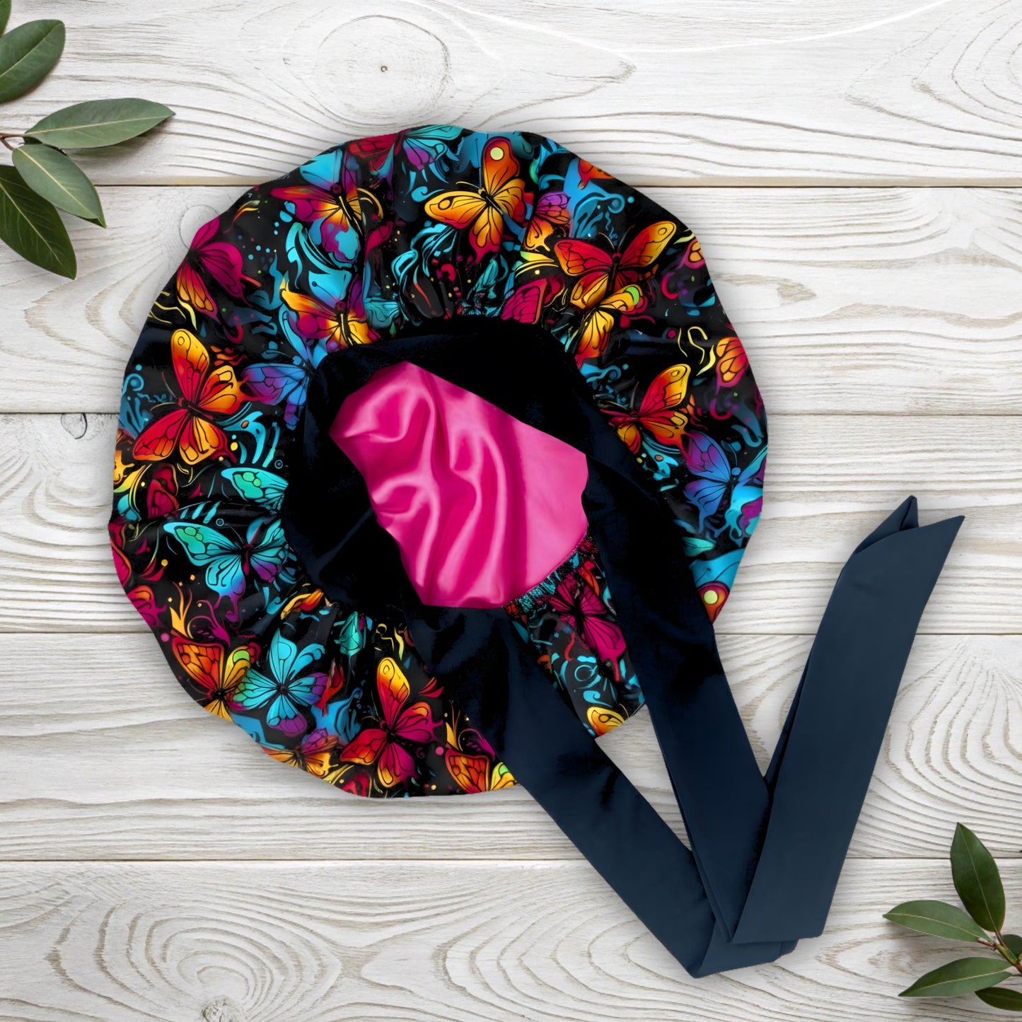 A colorful butterfly-patterned satin bonnet with a soft pink interior lining, displayed flat on a light wooden surface. The vibrant floral butterfly print and smooth charmeuse satin texture highlight its handmade quality. This silk sleep bonnet is designed to protect natural and curly hair from frizz and breakage while maintaining a stylish, elegant look. Perfect for luxury hair care by Prince Hair Company.