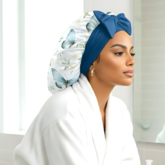 A woman wearing a handmade charmeuse silk satin hair bonnet featuring a floral butterfly print and long stretch ties tied into a bow, photographed in a bright, modern white bathroom. This satin sleep bonnet is designed to stay on all night, protect natural and curly hair, and reduce frizz. A luxury handmade silk bonnet by Prince Hair Company, perfect for overnight hair protection and style.