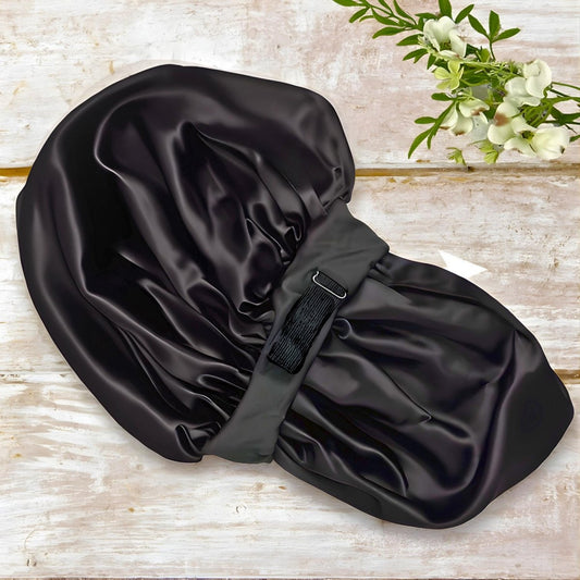 Black satin bonnet laid open on pale wooden boards, showing the wide band, elastic strap and sliding adjuster.