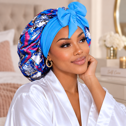 A woman wearing a bright blue satin bonnet with a large bow and a matching patterned bonnet underneath, paired with a white satin robe in a softly lit bedroom setting.
