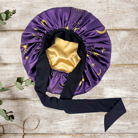 Blue Celestial Stretch Tie Satin Bonnet with gold charmeuse lining, featuring moon and star print on silky purple and blue fabric. Adjustable sleep cap for natural hair, braids, locs, and curly styles.
