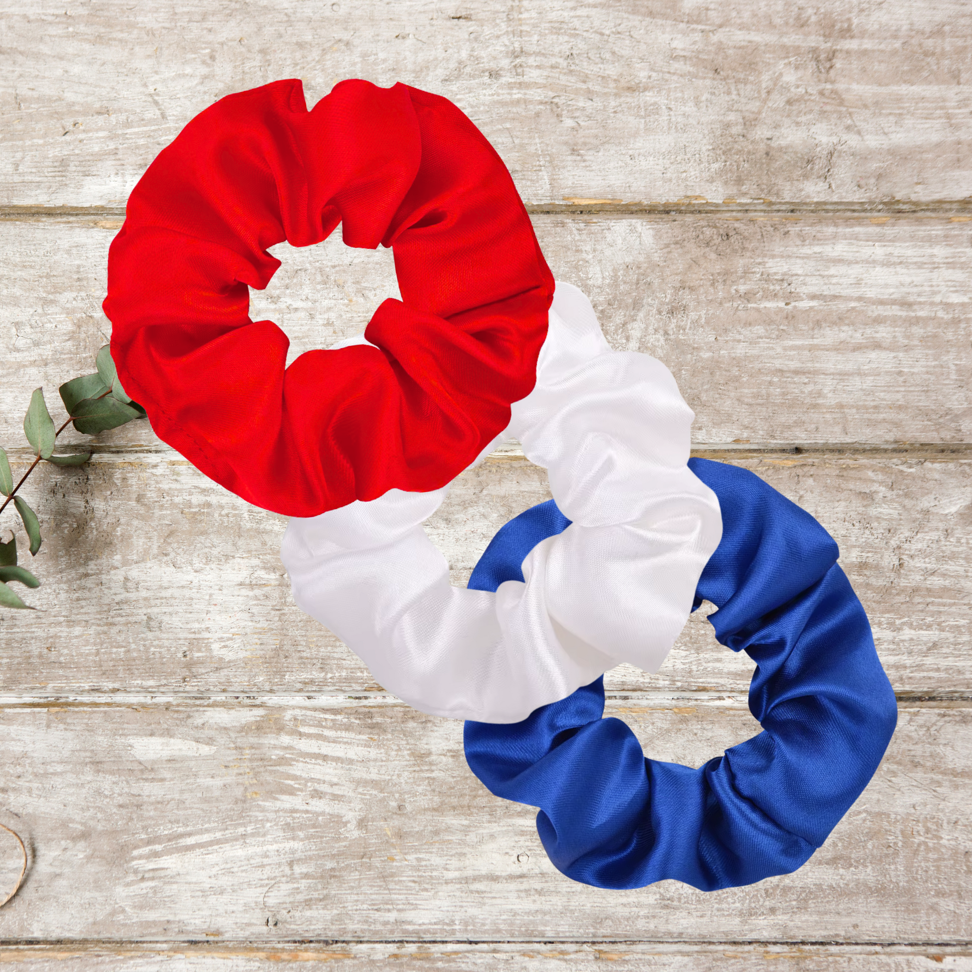Red, white, and blue satin scrunchie set arranged in a flat lay on a white wood background, perfect for patriotic hairstyles.