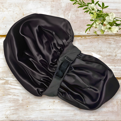 Prince Hair Company Black Charmeuse Silk satin hair bonnet on a wooden surface with a floral arrangement.