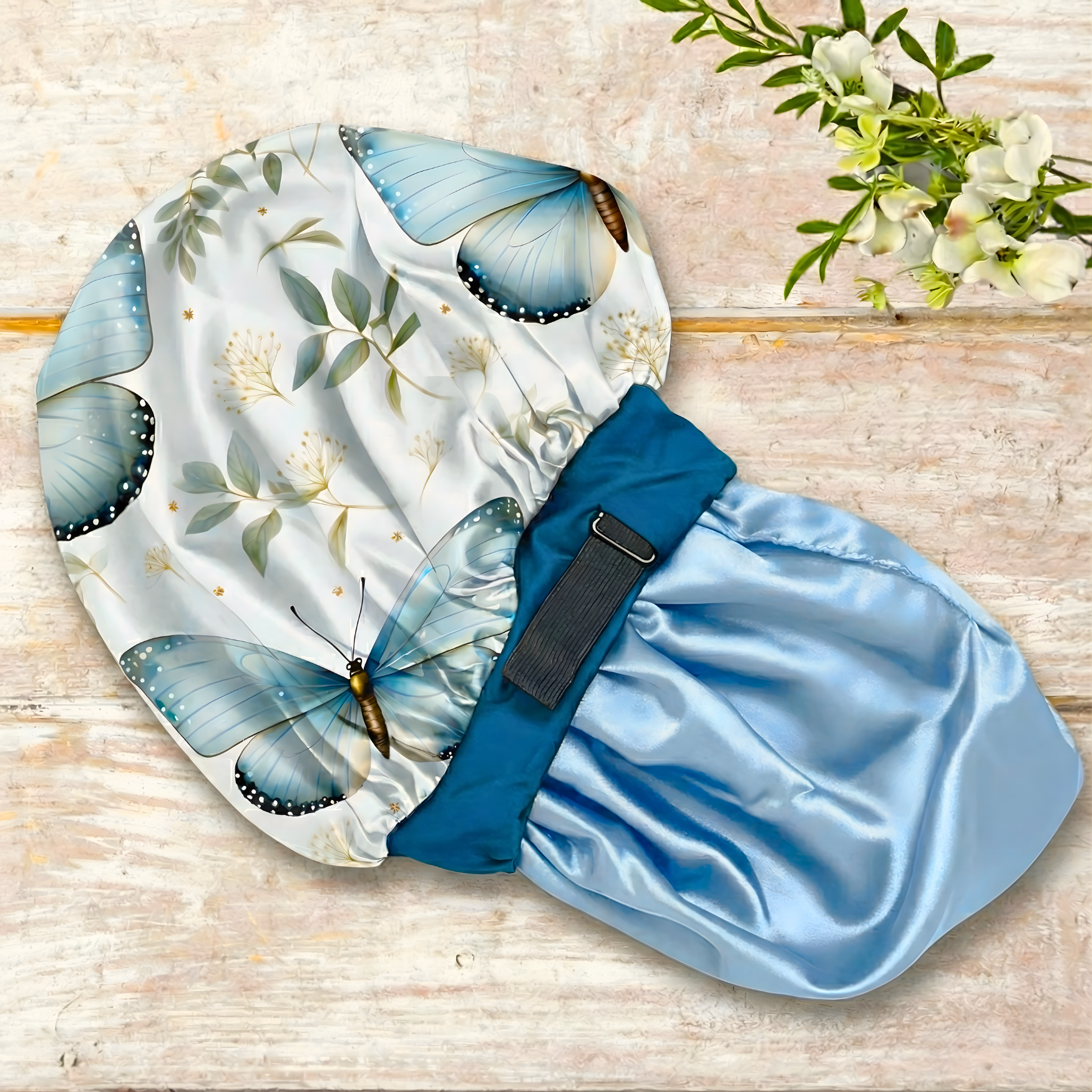 Display of a Charmeuse silk satin no-slip wide band hair bonnet in a floral butterfly print with powder blue silk lining and deep peacock blue band, showing a close-up of the adjustable elastic pull for a secure, edge-friendly fit, styled on a wooden table for protective nighttime hair care.