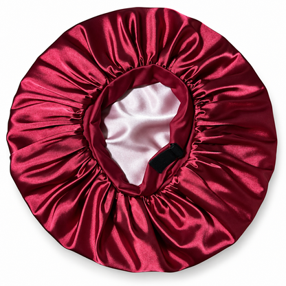 A satin sleep cap or bonnet in deep burgundy red, shown from a top-down view on a white background. The inner lining is a light pinkish-white satin, and a small black fabric tag is visible inside the opening.