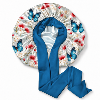 A satin sleep bonnet with a blue satin neck strap, featuring a white satin body printed with blue butterflies and red and white flowers, shown on a plain white background.