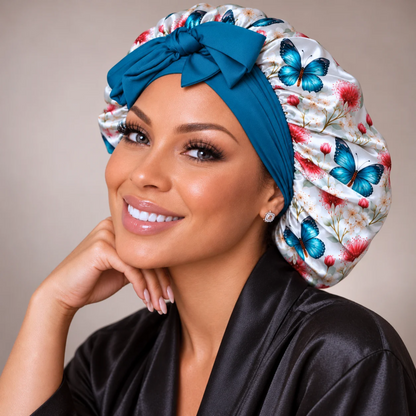 A smiling woman wearing a satin sleep cap with a large bow, featuring a white background with blue butterflies and red floral motifs, paired with a black satin robe against a soft beige background.