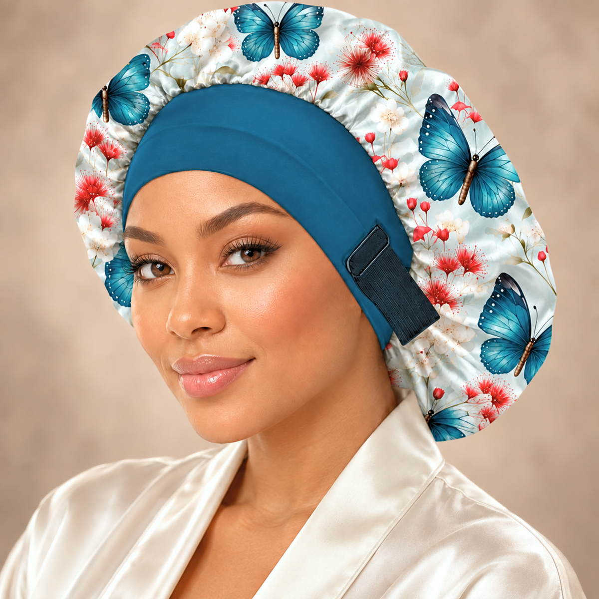 A woman wearing a satin sleep cap with a blue butterfly and red floral print on a white background, paired with a solid blue headband and a white satin robe against a soft beige studio background.