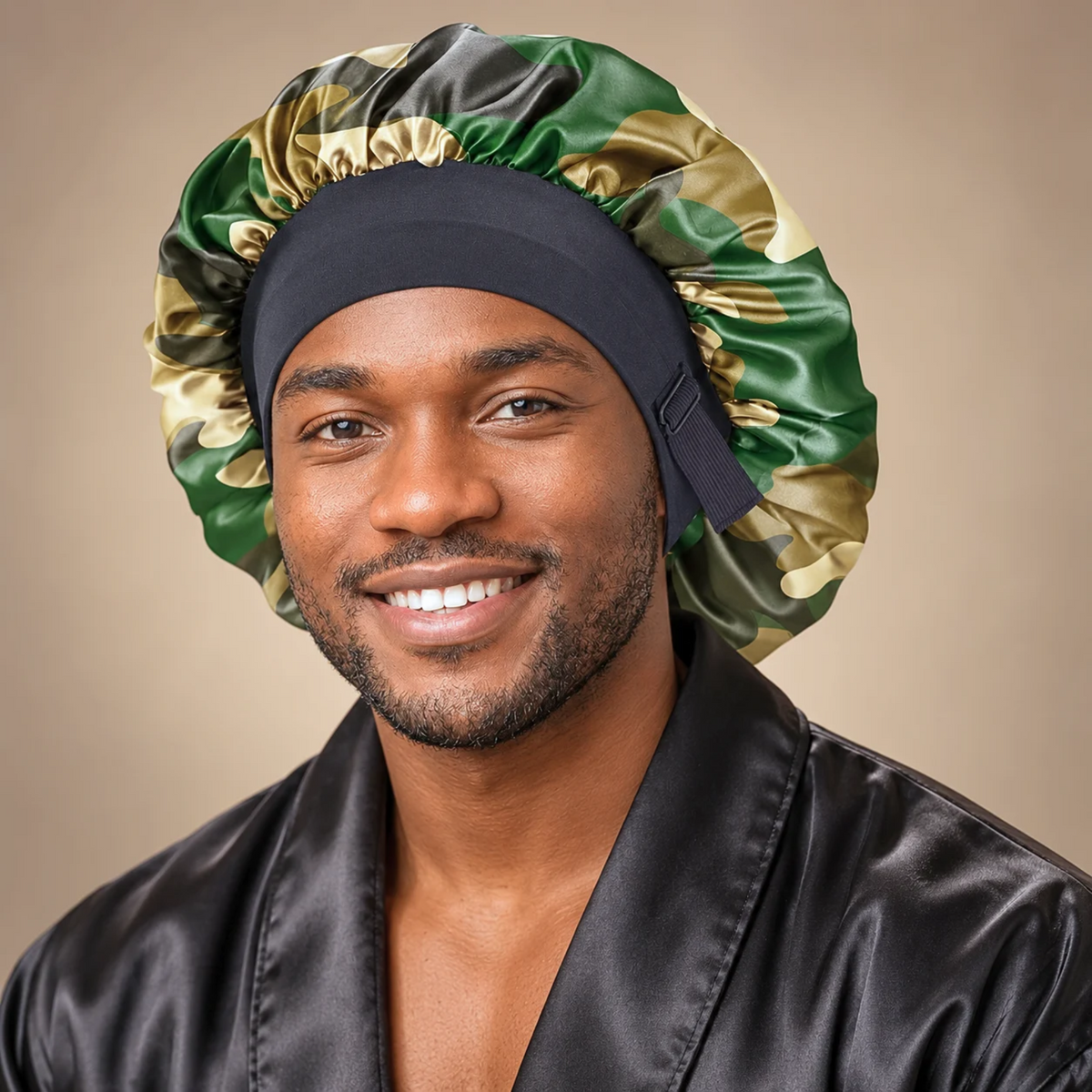 A smiling man wearing a shiny satin sleep cap with a green, gold, and black camouflage pattern, paired with a black satin robe against a warm beige background.