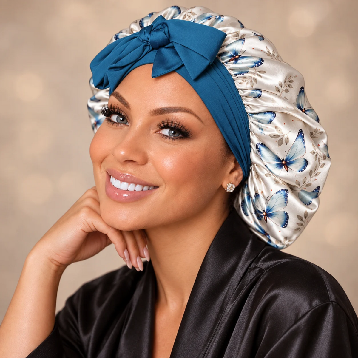 A smiling woman wearing a satin sleep cap with a large blue bow and a butterfly print, paired with a black satin robe against a soft beige background.