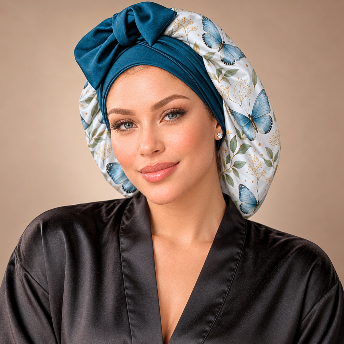 A woman wearing a satin sleep cap with a large bow and a blue butterfly floral print, paired with a black satin robe, set against a solid beige background.