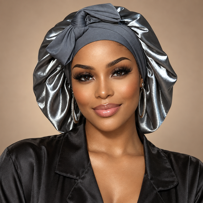 A woman wearing a shiny dark gray satin sleep cap with a large twisted top knot, paired with a black satin robe, set against a solid beige background.