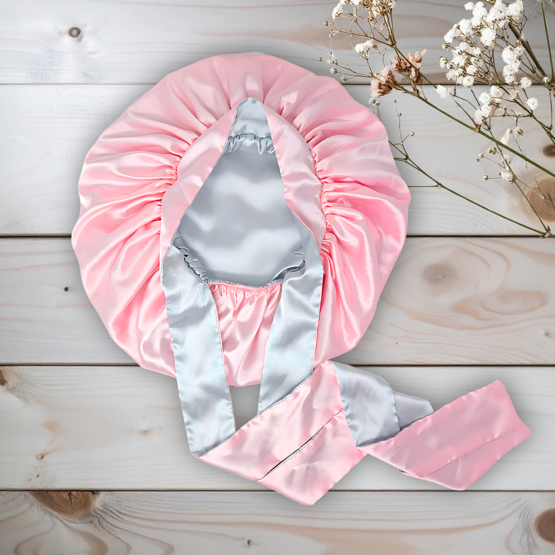 Pink and gray reversible all satin hair bonnet on a wooden surface with white flowers.