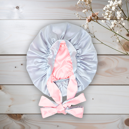 Beautiful Gray  satin bonnet with pink interior and bow on a wooden surface with white flowers.