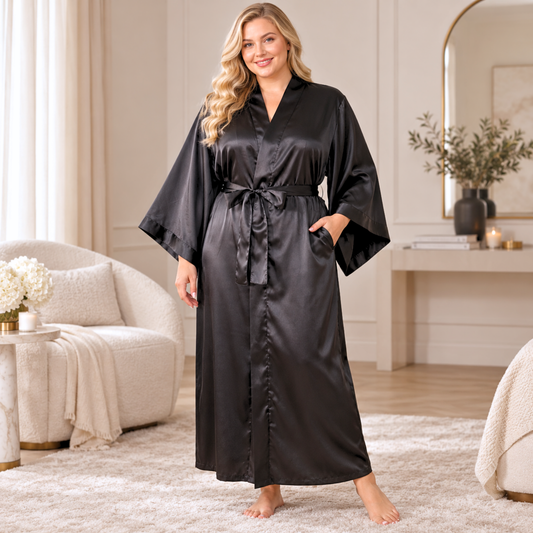 A plus size woman stands barefoot in a softly lit bedroom or spa-like interior, wearing a long black satin robe with wide sleeves and a self-tie belt. The robe has a smooth, slightly shiny finish and side pockets.