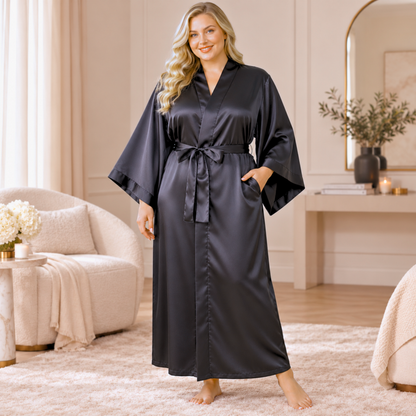 A woman modeling a long black satin robe in a bright, modern bedroom setting. The robe features wide sleeves, a matching tie belt, and side pockets.
