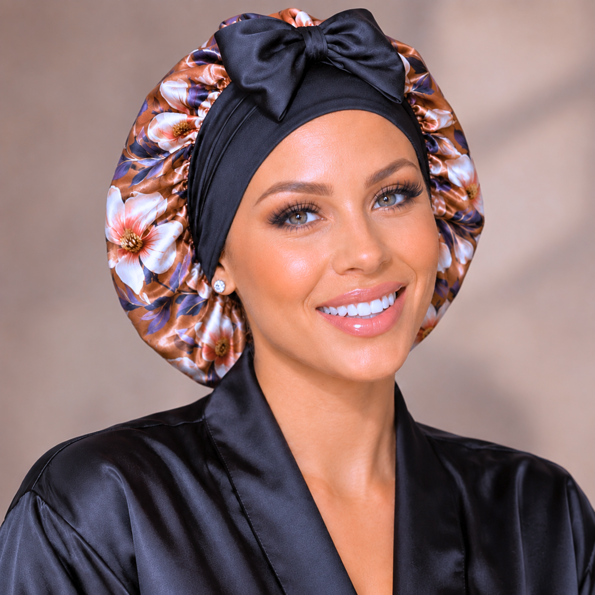 A smiling woman wearing a black satin bonnet with a large bow and a matching floral satin robe. The bonnet and robe feature a brown, gold, and white floral pattern, shown against a soft beige background.