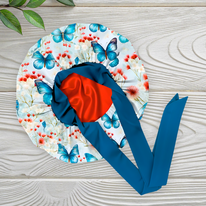 A handmade charmeuse satin stretch tie bonnet displayed flat on a light wooden surface. The bonnet features a blue butterfly and floral pattern with red and white flowers, a deep blue band with long adjustable ties, and a bright red satin interior lining that adds a pop of color and luxury.