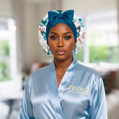A woman wearing a light blue satin robe with “Prince Hair Company” in gold lettering poses confidently against a blurred background. She wears a blue butterfly-print charmeuse satin bonnet with a matching blue stretch tie and bow, showcasing a polished and elegant look that highlights the bonnet’s luxurious design and fit.
