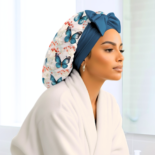 A beautiful woman wearing a butterfly print charmeuse satin hair bonnet with blue stretch ties and bow sits at a bright bathroom vanity in a white robe. The handmade satin sleep cap from Prince Hair Company protects natural, curly, and protective hairstyles while adding luxury to her self-care routine. Perfect example of a silk satin bonnet for hair protection and beauty sleep.