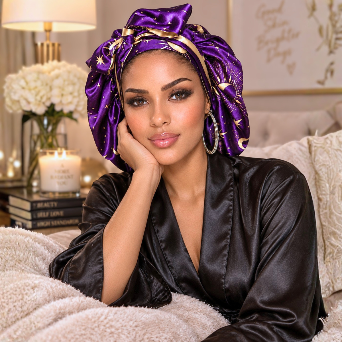 A woman wearing a shiny purple satin turban headwrap with gold star details and a matching black satin robe, posing on a cozy sofa with a blanket.