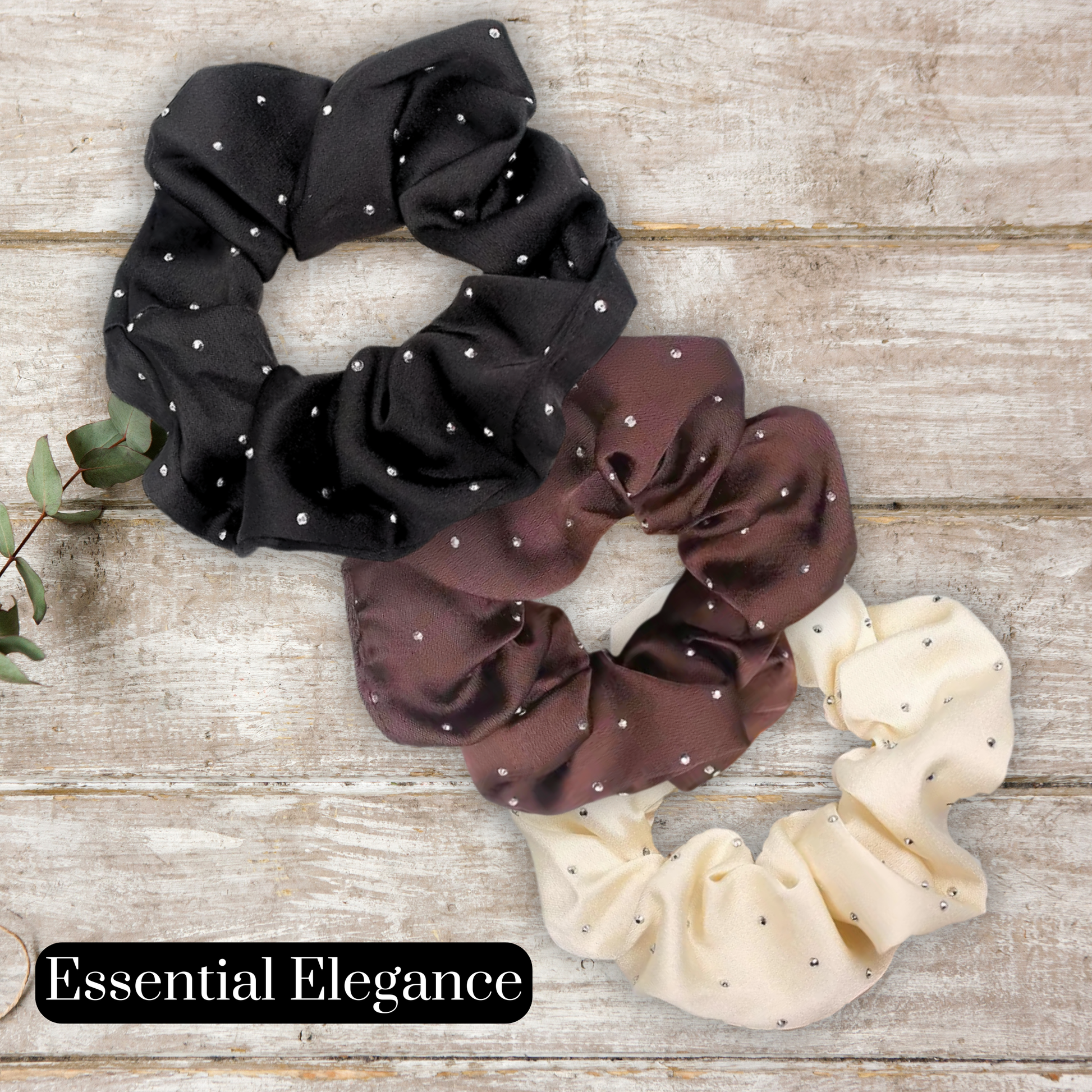 Three satin rhinestone scrunchies in black, brown, and beige on a wooden surface with 'Essential Elegance' branding.