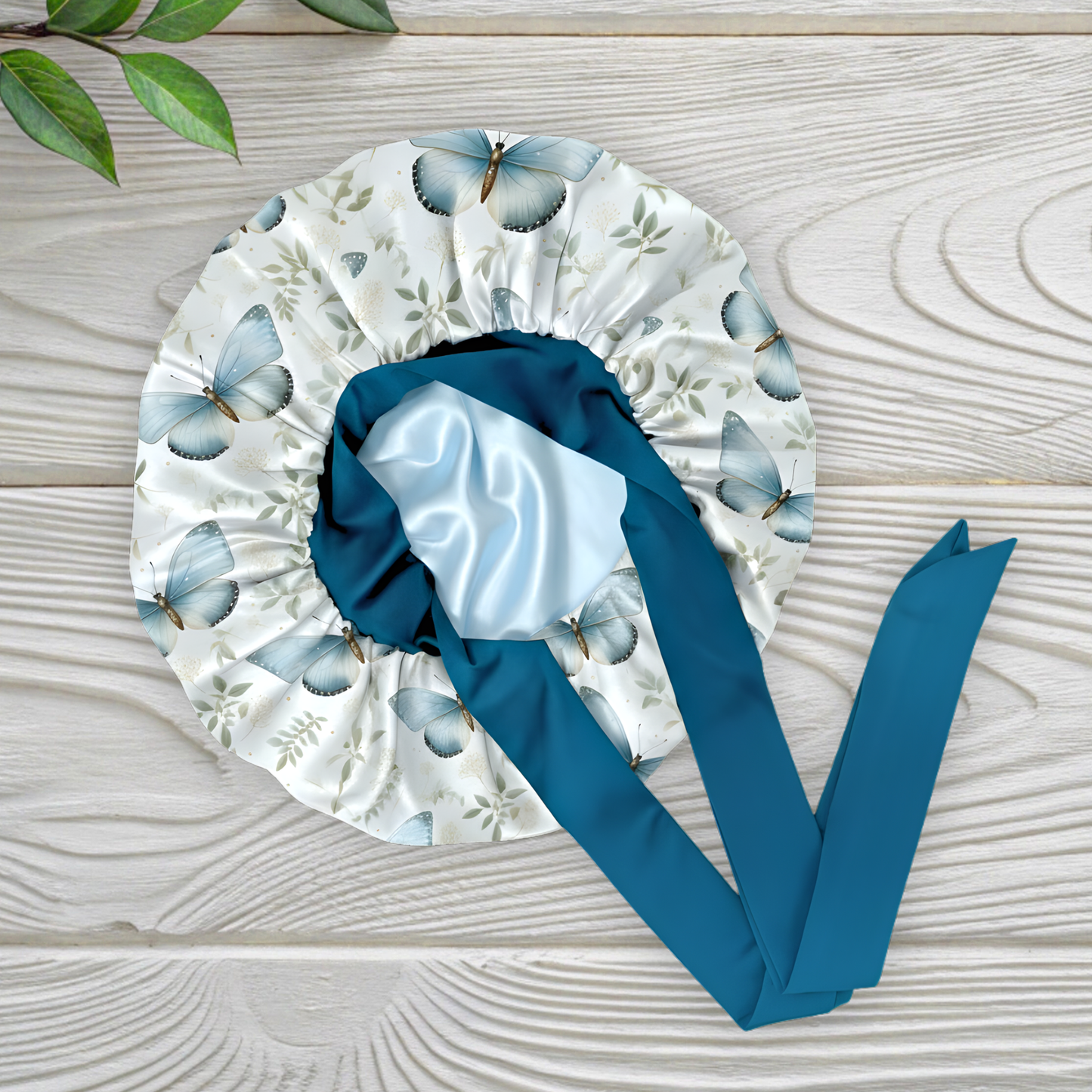 A butterfly floral-patterned charmeuse satin silk hair bonnet with long peacock-blue stretch ties tied into a bow, displayed flat on a light wooden background with a small green plant in the corner. This luxury handmade satin sleep bonnet is crafted for natural and curly hair protection, reducing frizz and dryness while staying stylish. Designed and handmade by Prince Hair Company in Chicago.