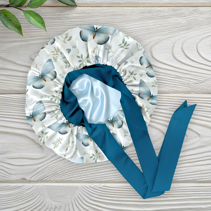 A butterfly floral-patterned charmeuse satin silk hair bonnet with long peacock-blue stretch ties tied into a bow, displayed flat on a light wooden background with a small green plant in the corner. This luxury handmade satin sleep bonnet is crafted for natural and curly hair protection, reducing frizz and dryness while staying stylish. Designed and handmade by Prince Hair Company in Chicago.