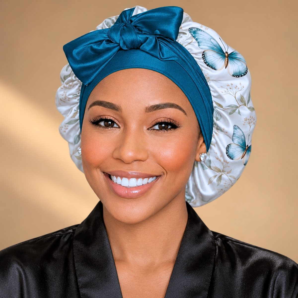 A smiling woman wearing a satin bonnet with a large teal bow and white side panels printed with blue butterflies, paired with a black satin robe against a warm beige background.