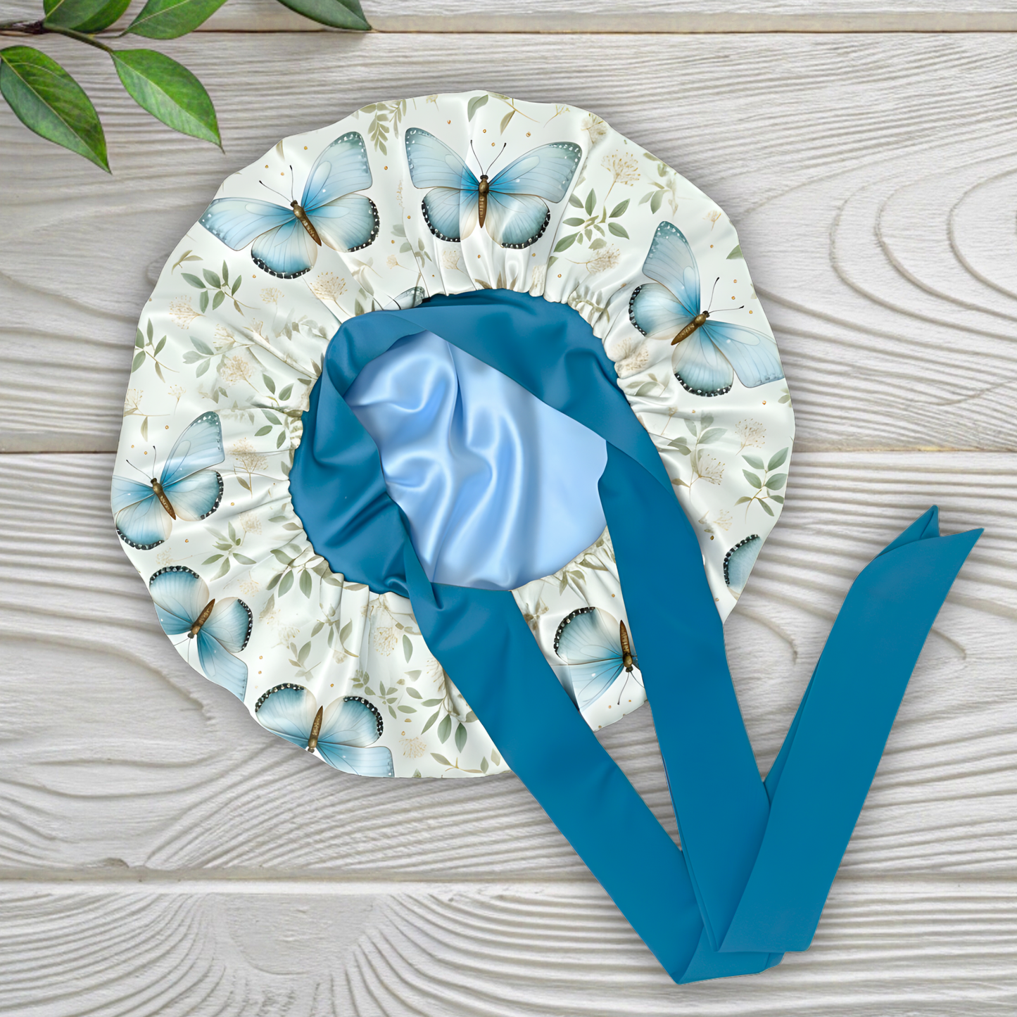 Charmeuse satin silk hair bonnet with a blue butterflies surrounded by a floral print with long peacock blue stretch ties on a white wooden surface.