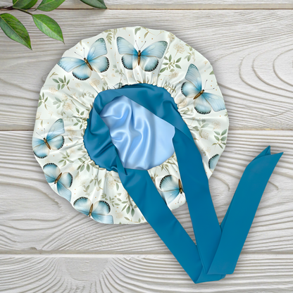 Charmeuse satin silk hair bonnet with a blue butterflies surrounded by a floral print with long peacock blue stretch ties on a white wooden surface.