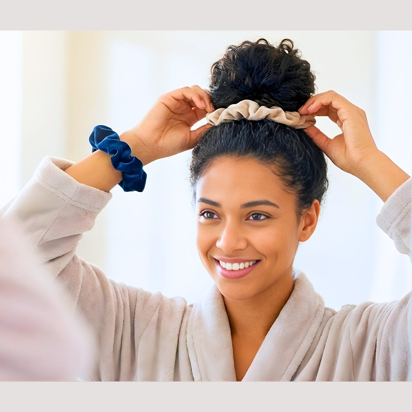 A smiling biracial woman with dark curly hair wearing a light plush robe looks into a mirror while styling her hair into a high bun. She is using a Latte beige velvet scrunchie to secure her bun and is wearing a Prussian blue velvet scrunchie on her wrist, demonstrating versatile and damage-free hair styling.