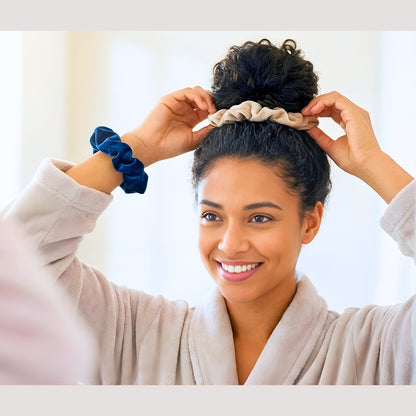 A smiling biracial woman with dark curly hair wearing a light plush robe looks into a mirror while styling her hair into a high bun. She is using a Latte beige velvet scrunchie to secure her bun and is wearing a Prussian blue velvet scrunchie on her wrist, demonstrating versatile and damage-free hair styling.
