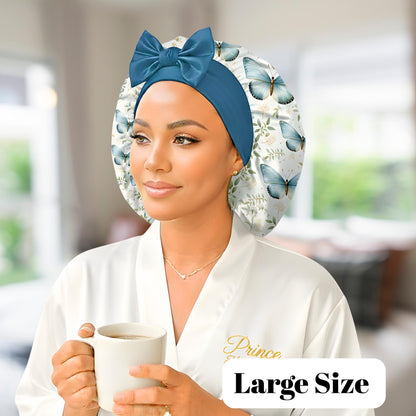 A woman wearing a butterfly floral-patterned charmeuse satin silk hair bonnet with long stretch ties formed into a bow, holding a mug in her hands. She is photographed in a softly lit indoor setting with a blurred background, highlighting the bonnet’s elegant shine and handmade quality. This luxury satin sleep cap protects natural, curly, and textured hair while maintaining a cute, comfortable fit.