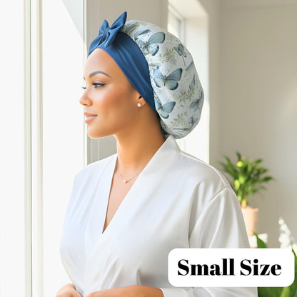 Side profile of a woman wearing a white robe and a handmade charmeuse silk hair bonnet with a floral butterfly print, gazing thoughtfully into the distance. The small satin bonnet fits comfortably on short to mid-back length hair—straight, curly, wav