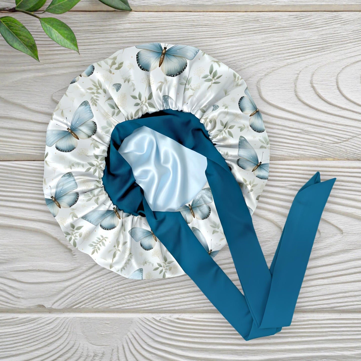 A butterfly floral-patterned charmeuse satin silk hair bonnet with long peacock-blue stretch ties tied into a bow, displayed flat on a light wooden background with a small green plant in the corner. This luxury handmade satin sleep bonnet is crafted for natural and curly hair protection, reducing frizz and dryness while staying stylish. Designed and handmade by Prince Hair Company in Chicago.