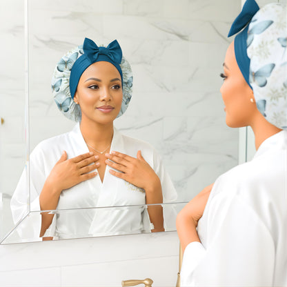 Side profile of a woman wearing a butterfly floral-patterned charmeuse satin silk hair bonnet with long peacock blue stretch ties tied into a neat bow, looking into a mirror. The soft lighting highlights the bonnet’s silky sheen and handmade quality. This luxury satin sleep bonnet is designed to protect natural, curly, and textured hair from frizz, dryness, and breakage while maintaining a stylish, elegant look.