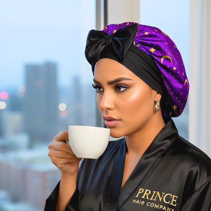 Model drinking coffee by a window wearing a black Prince Hair Company robe and the Celestial Plum satin bonnet.