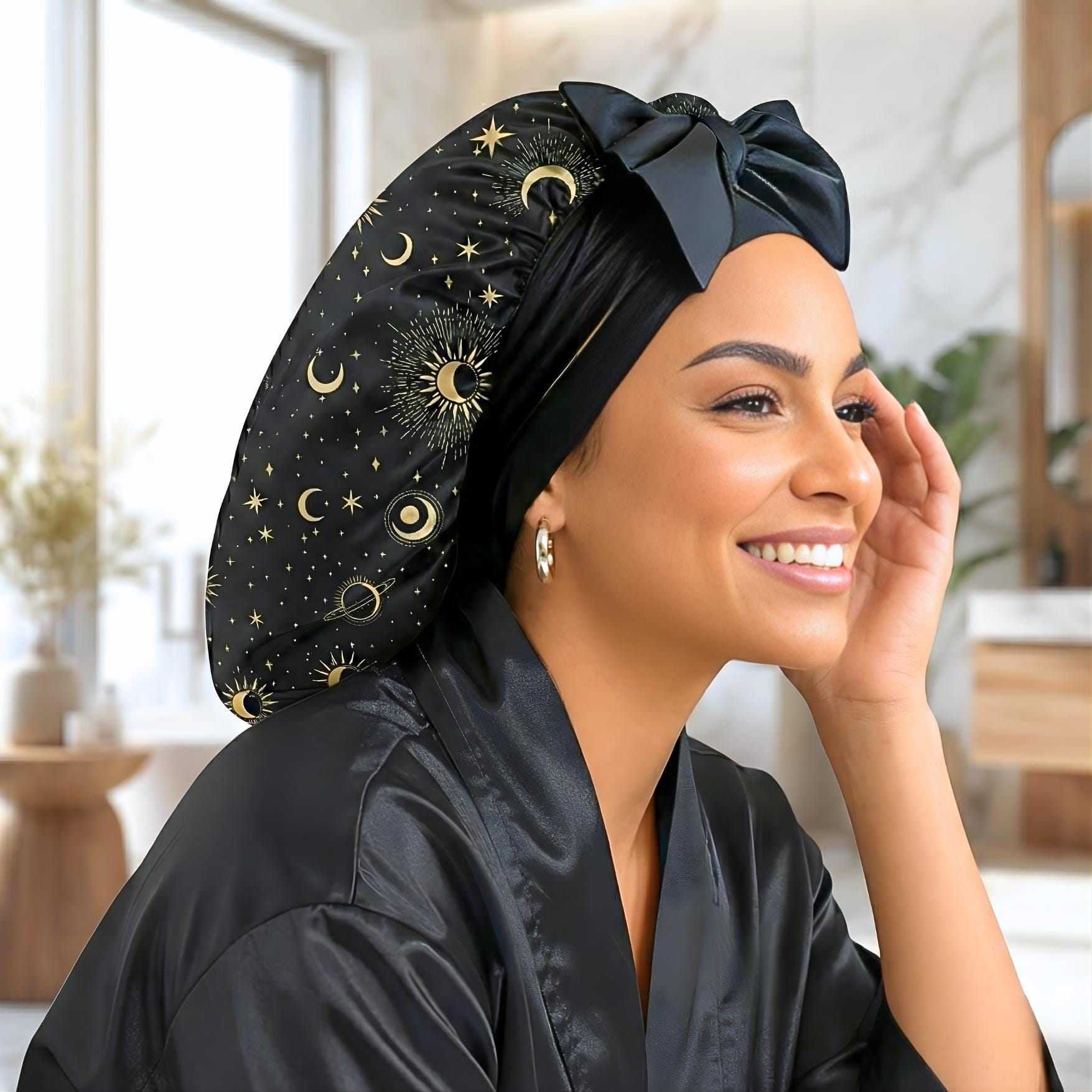 Close-up of a smiling model wearing the Starry Night satin bonnet. She has a radiant, happy expression and looks off-camera. The bonnet is black with a gold celestial print of moons, stars, and sunbursts, featuring a large, elegant bow at the front. She wears a matching black satin robe with "Prince Hair Company" embroidered in gold on the lapel. The background is a bright, soft-focus modern bathroom or bedroom setting