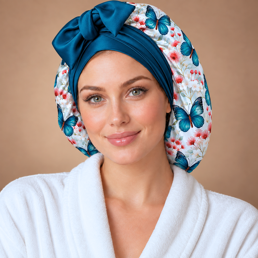 A woman wearing a white bathrobe and a satin turban-style hair wrap with a blue floral and butterfly print, posed against a warm beige studio background.