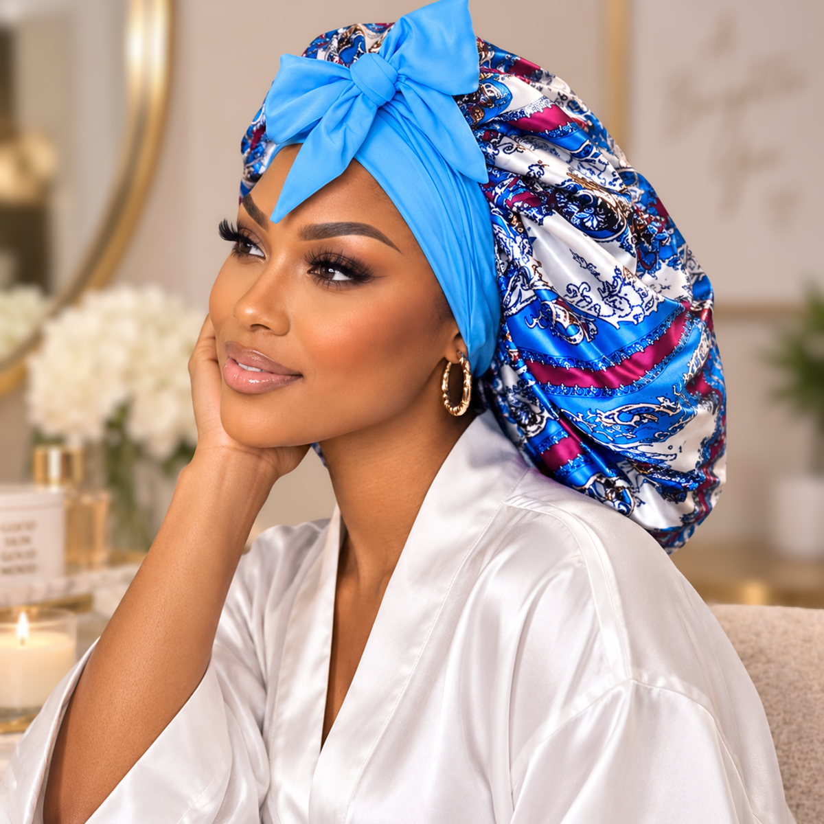 A woman wearing a shiny blue satin bonnet with a large bow and a matching blue, pink, and white patterned hair scarf, paired with a white satin robe in a softly lit indoor setting.