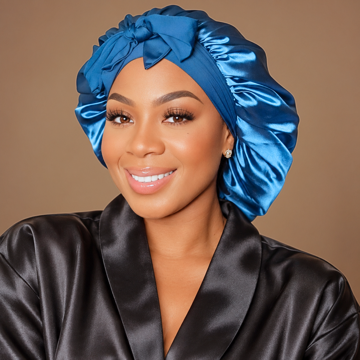 A woman wearing a shiny peacock blue satin bonnet with a large bow and a black satin robe, set against a solid warm brown background.