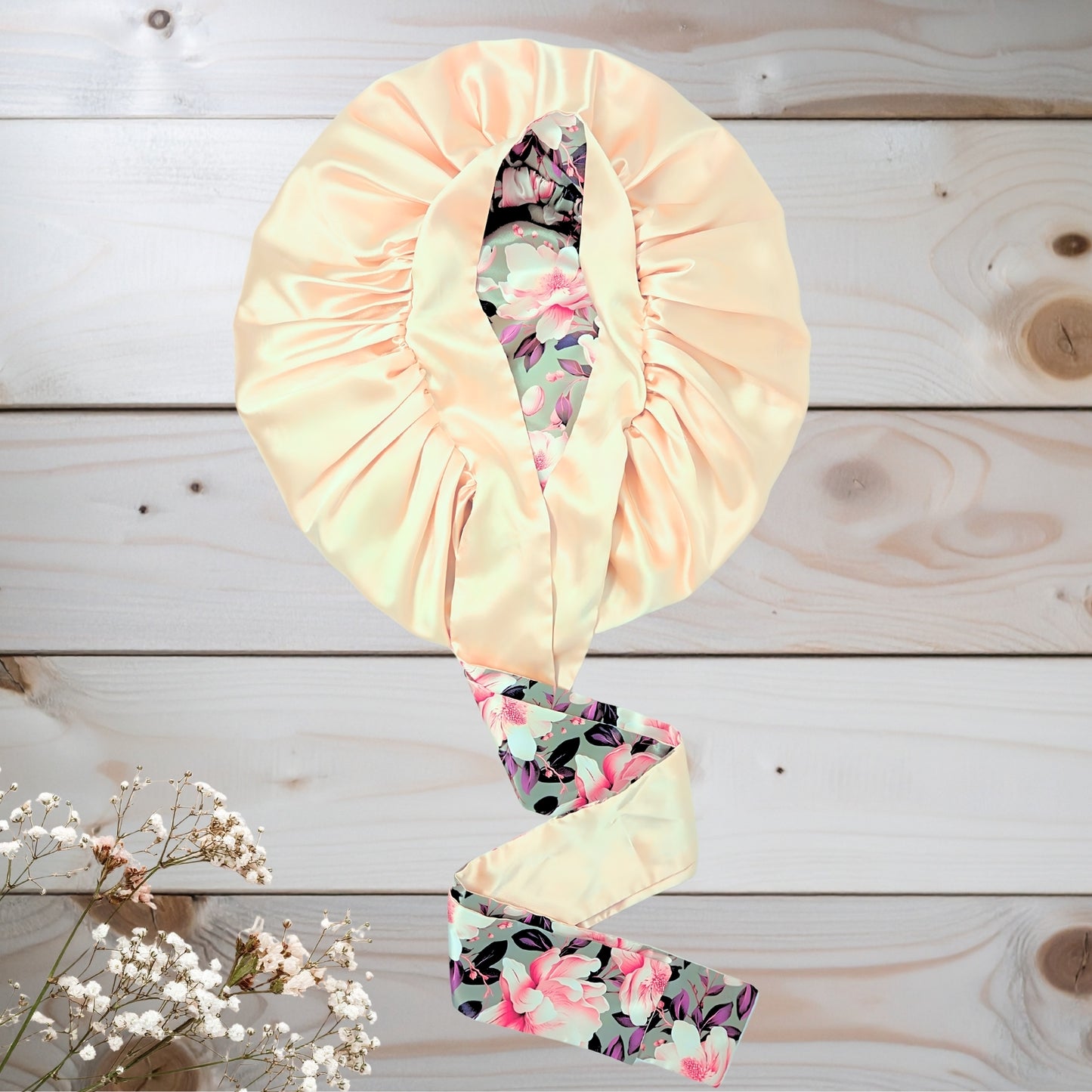 A reversible handmade satin sleep bonnet laid flat on a light wooden surface next to white baby's breath flowers. The exterior is a solid champagne yellow color, slightly open to reveal the inner lining featuring a green, pink, and white Olive Magnolia floral print. It includes long, stretch-free satin ties.