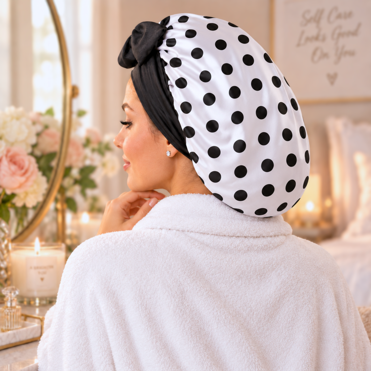 A woman in a white robe shown from behind wearing a white satin-like hair bonnet with black polka dots and a black bow, in a softly lit bedroom setting.