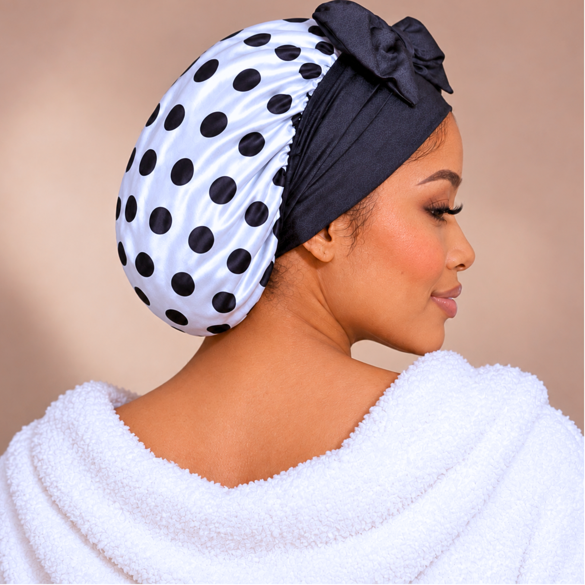 A woman shown in profile wearing a white satin bonnet with black polka dots and a black bow headband, with a white towel draped over her shoulders against a soft beige background.