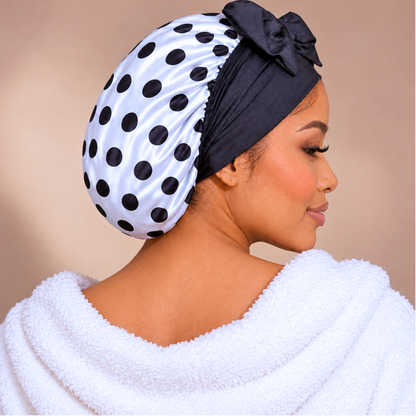 A woman shown in profile wearing a white satin bonnet with black polka dots and a black bow headband, with a white towel draped over her shoulders against a soft beige background.